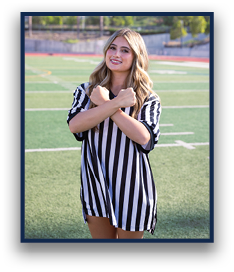 A person with long wavy hair wearing a black and white striped referee shirt stands on a sports field with arms crossed in front of their chest. Description generated by AI