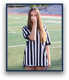 A person with long straight hair wearing a black and white striped shirt stands on a sports field with one hand partially covering their face. Description generated by AI