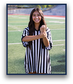 A person with long hair wearing a black and white striped referee shirt is smiling and holding one arm with the other on a sports field. Description generated by AI