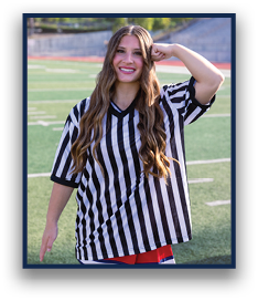 A person with long wavy hair wearing a black and white striped shirt and a colorful skirt is standing on a sports field smiling and posing with one arm bent and fist near their head. Description generated by AI