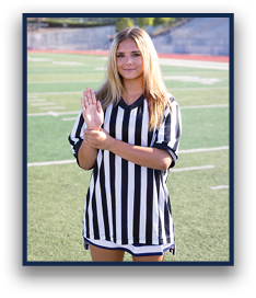 A person wearing a black and white striped referee shirt is standing on a sports field holding one hand up with the other hand gripping the wrist. Description generated by AI