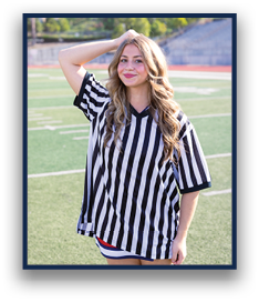 A person with long wavy hair wearing a black and white striped referee shirt stands on a sports field with one hand resting on their head and a slight smile. Description generated by AI