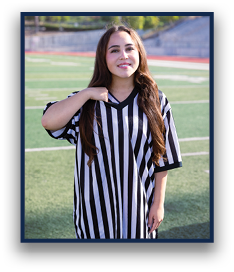 A person with long hair wearing a black and white striped referee shirt stands on a sports field with a slight smile. Description generated by AI