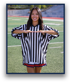 A person with long hair wearing a black and white striped referee shirt is standing on a sports field smiling and holding their hands parallel in front of them. Description generated by AI