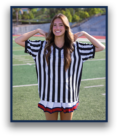 A person with long wavy hair is smiling and posing with hands behind their head while wearing a black and white striped referee shirt and a pleated skirt on a sports field. Description generated by AI