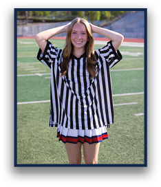 A person with long hair wearing a black and white striped referee shirt and a pleated skirt stands on a sports field with hands resting on their head, smiling. Description generated by AI