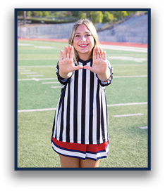 A person wearing a black and white striped referee shirt and a red, white, and blue skirt is standing on a sports field smiling and holding their hands up with palms facing forward. Description generated by AI
