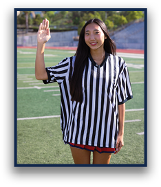 A person with long hair wearing a black and white striped referee shirt is standing on a sports field with one hand raised. Description generated by AI