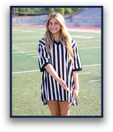 A person with long hair wearing a black and white striped referee shirt is standing on a sports field smiling and gesturing with one hand. Description generated by AI