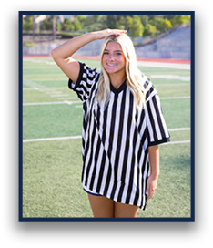 A person with long light hair wearing a black and white striped referee shirt stands on a sports field with one hand on their head and a slight smile. Description generated by AI