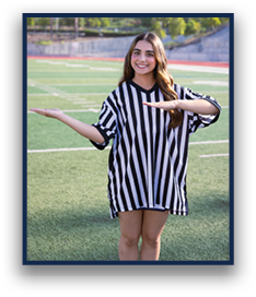A person with long hair wearing a black and white striped shirt is standing on a sports field smiling and holding their arms out horizontally at different heights. Description generated by AI
