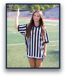 A person with long hair wearing a black and white striped referee shirt is standing on a sports field with one arm raised. Description generated by AI