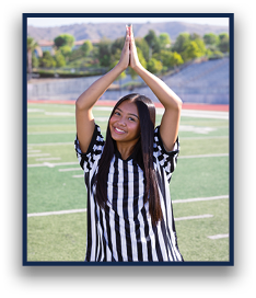 A person wearing a black and white striped referee shirt is standing on a sports field with hands pressed together above their head and smiling. Description generated by AI
