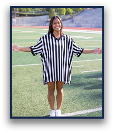 A person wearing a black and white striped referee shirt stands on a sports field with arms extended and smiling. Description generated by AI