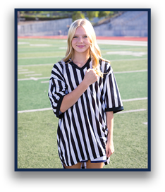 A person with long hair wearing a black and white striped referee shirt stands on a sports field with a slight smile and one hand raised to their chest. Description generated by AI