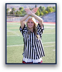 A person with long hair wearing a black and white striped referee shirt is standing on a sports field with arms crossed above their head and smiling. Description generated by AI