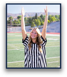 A person wearing a black and white striped referee shirt is standing on a sports field with arms raised and smiling. Description generated by AI