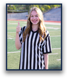A person with long hair wearing a black and white striped shirt is standing on a sports field smiling and giving a thumbs up. Description generated by AI