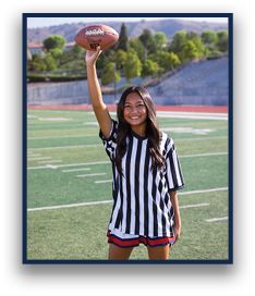 A person with long hair wearing a black and white striped shirt and red shorts is standing on a football field smiling and holding a football up in one hand. Description generated by AI