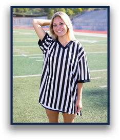 A person with long light hair is smiling and standing on a sports field wearing a black and white vertically striped oversized shirt with one arm raised behind their head. Description generated by AI