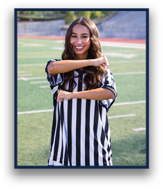 A person with long wavy hair wearing a black and white striped shirt is smiling and crossing their arms in front of their chest while standing on a sports field. Description generated by AI