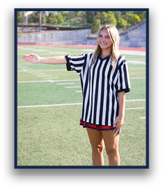 A person wearing a black and white striped referee shirt stands on a sports field with one arm extended to the side and is smiling. Description generated by AI