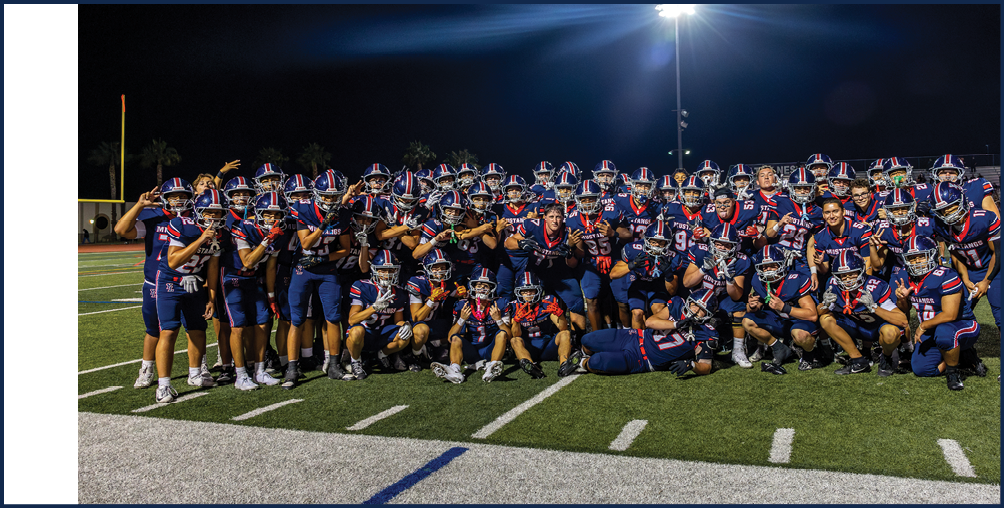 A large group of people wearing matching football uniforms pose together on a football field at night under bright stadium lights. Description generated by AI