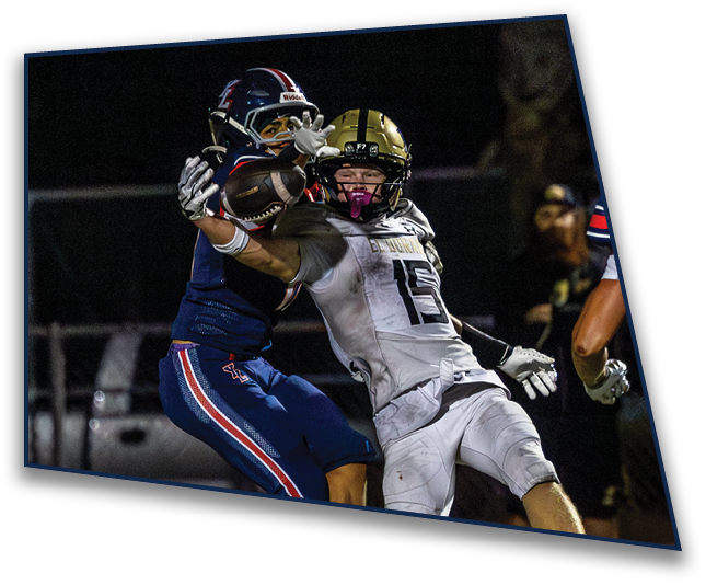 Two people in football gear are reaching out to catch a football during a nighttime game. Description generated by AI