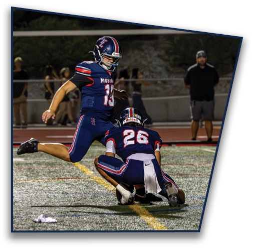 Two people in football uniforms are on a field at night with one holding the ball on the ground and the other kicking it. Description generated by AI