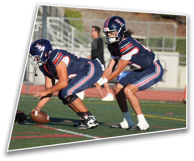 Two people wearing football helmets and uniforms are positioned on a field with a blurred background of a fence, stairs, and greenery. Description generated by AI