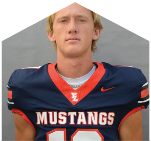 A person with short light hair wearing a navy blue football jersey with red and white accents and the word Mustangs across the chest stands against a plain gray background. Description generated by AI