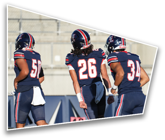 Two people wearing navy blue football uniforms and helmets with red and white stripes are seen from behind on a field. Description generated by AI