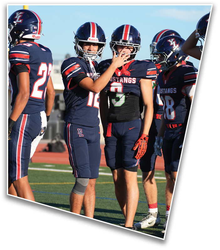 Four people in football uniforms and helmets stand on a field with one person gesturing with their hand near another's face. Description generated by AI