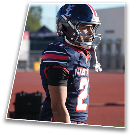A person wearing a navy blue football uniform with red and white stripes and a helmet stands on a sports field looking to the side. Description generated by AI