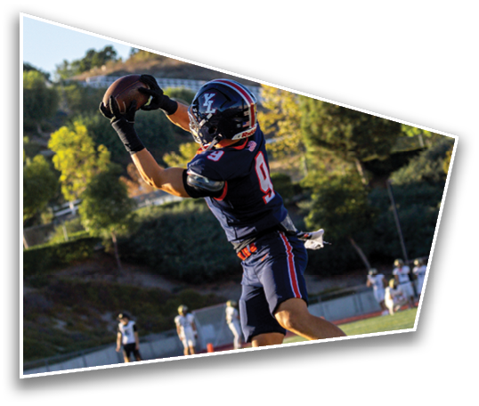 A person in a dark sports uniform is jumping or reaching up on a football field with other players and trees in the background. Description generated by AI