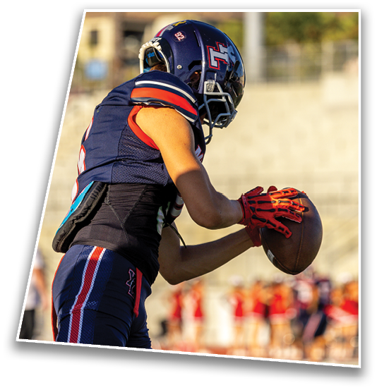 A person wearing a blue and red sports uniform and red gloves is holding a football in a ready-to-throw position on a field with blurred spectators in the background. Description generated by AI