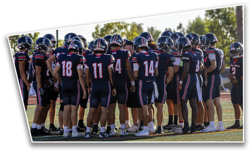 A group of people wearing matching dark sports uniforms and helmets are huddled together on a field during daylight. Description generated by AI