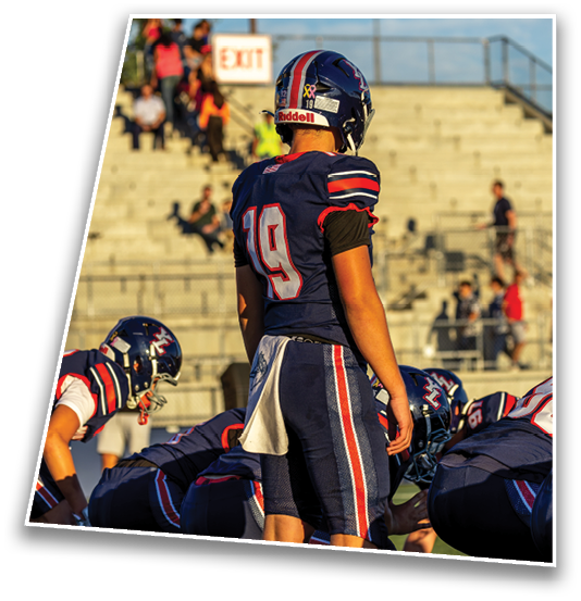 A group of people in football uniforms are positioned on a field with one standing and others crouched, preparing for a play during a game. Description generated by AI