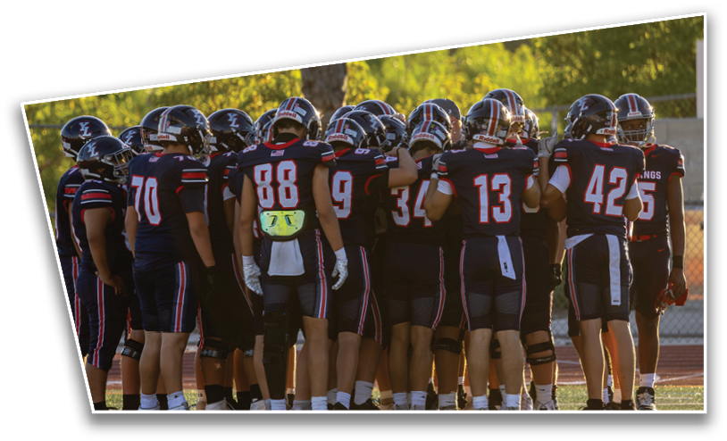 A group of people wearing dark sports uniforms with helmets are huddled together on a field, likely preparing for or discussing a football game. Description generated by AI