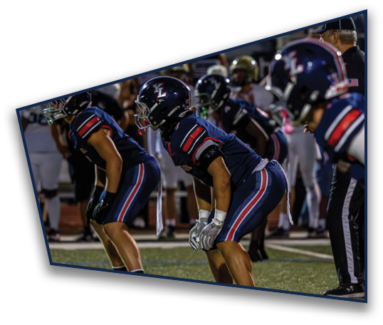 Several people in dark blue sports uniforms with helmets are crouched in a ready position on a football field during a game. Description generated by AI