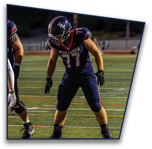 Two people in football uniforms are on a field with one holding a football while others stand in the background. Description generated by AI