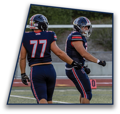Two people wearing navy blue football uniforms and helmets stand on a field with greenery in the background. Description generated by AI