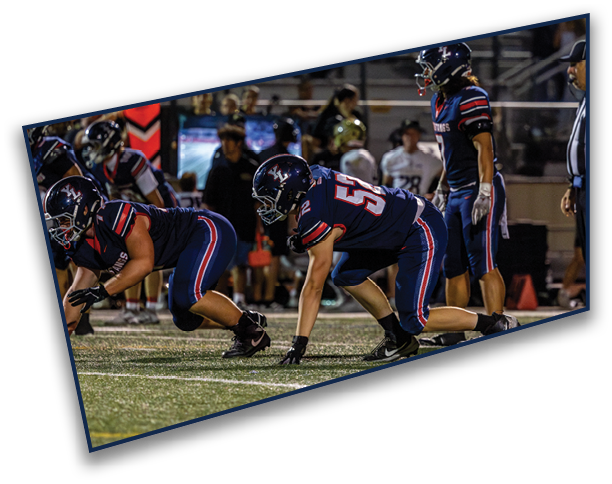 Three people in football uniforms are positioned on a field in a ready stance, preparing for a play during a nighttime game. Description generated by AI