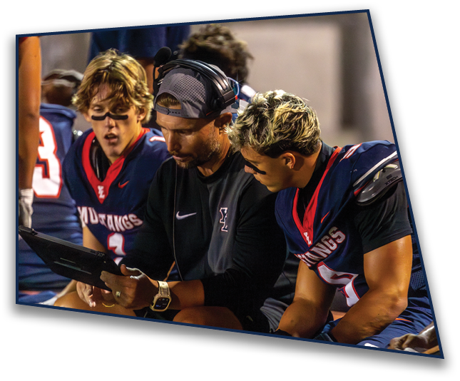 Three people in sports uniforms and a coach wearing a headset are closely looking at a tablet during a game. Description generated by AI
