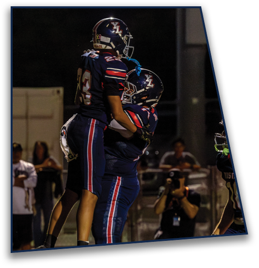 Two football players in dark uniforms with helmets celebrate on the field at night while people watch and a photographer captures the moment. Description generated by AI