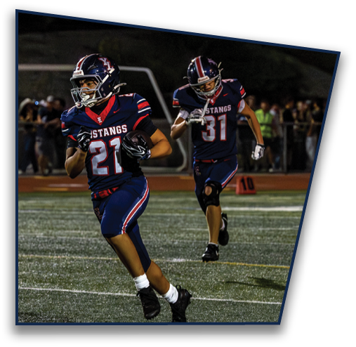 Two people in football uniforms with helmets are running on a field, with one holding a football and the other following behind. Description generated by AI
