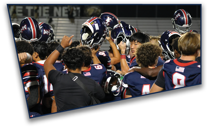 A group of people wearing football uniforms and helmets are gathered closely together on a field, appearing to celebrate or strategize. Description generated by AI