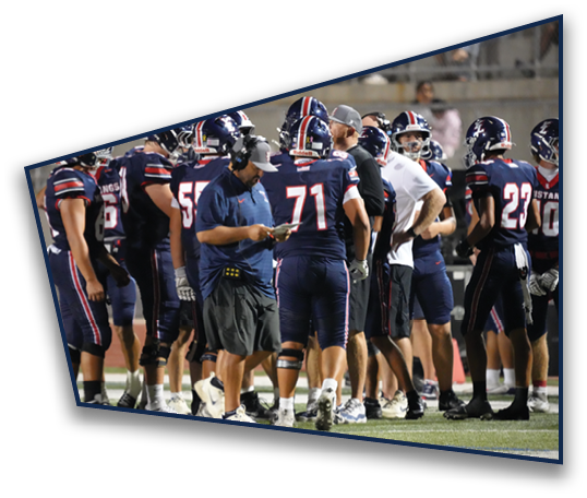 A group of people wearing football uniforms and helmets are gathered on a field with two adults in casual sportswear appearing to coach or discuss strategy. Description generated by AI