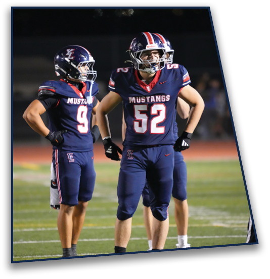 Three people in football uniforms stand on a field at night, with two wearing navy blue jerseys labeled “Mustangs" and one in black pants and a black shirt. Description generated by AI
