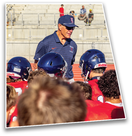 A coach wearing a navy blue cap and polo shirt is speaking to a group of people in red sports uniforms and navy blue helmets on a sports field. Description generated by AI